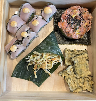 Une boîte en bois contenant des rouleaux de sushi avec de la sauce, un tartare de saumon sur des algues, une salade enveloppée dans une feuille et des craquelins croustillants aux algues.