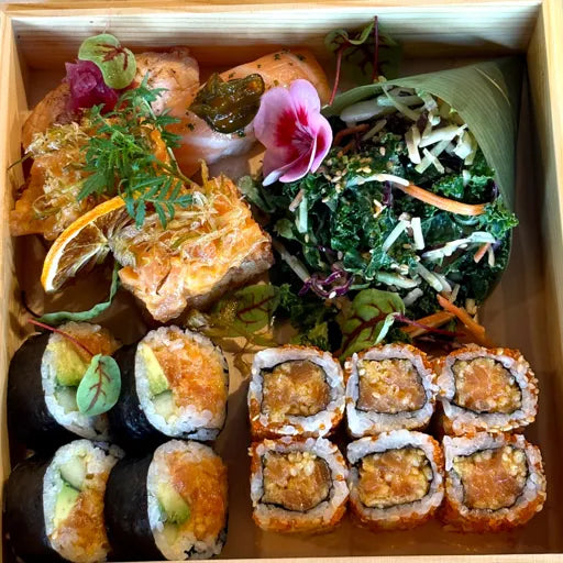 Une boîte en bois remplie de sushis, dont des rouleaux au saumon et à l'avocat, et des rouleaux au thon épicé. Elle contient également des bouchées de saumon, une salade enveloppée dans une feuille et une tranche d'orange séchée garnie.