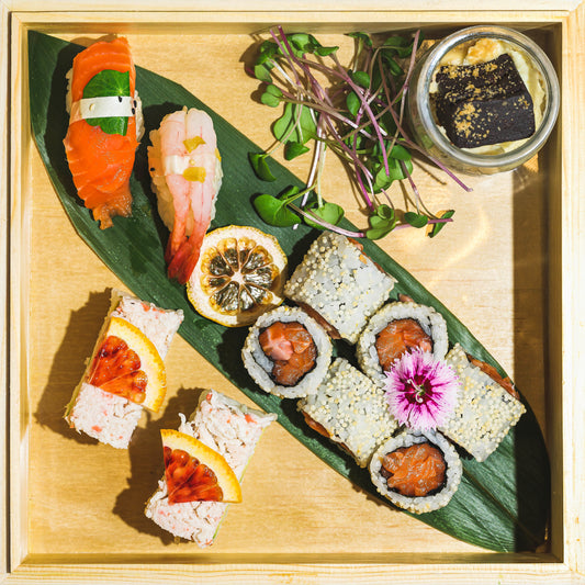 Une Kidzbox du Fish Club privé avec un assortiment de rouleaux de sushi, des nigiri, des micropousses et un petit pot de sauce soja sur une garniture de feuilles vertes. Les rouleaux comprennent du poisson cru, des fruits de mer et des garnitures. Fleur décorative incluse pour une touche d'élégance. Parfait à emporter dans les meilleurs spots de sushi de la Ville de Québec.