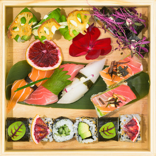 Un plateau en bois présentant un assortiment de sushis, dont des nigiri, des sashimis et des makis, garni de tranches d'agrumes, de fleurs comestibles et de verdure, parfait pour emporter à Ville de Québec. La Fishbox Végé de FISH est un choix idéal pour tout amateur de sushi.
