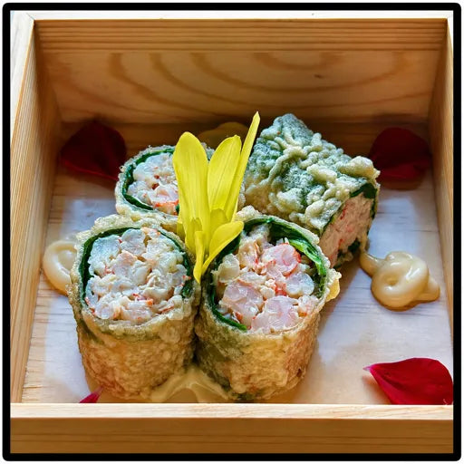 Trois rouleaux de sushi Rouleau de matanes croustillantes de FISH, surmontés d'une fleur jaune, servis dans une boîte en bois avec des cuillerées de mayonnaise et des pétales de fleurs rouges.