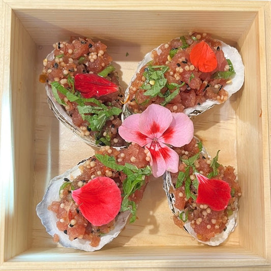 Tartare & Coquillages by FISH présente un tartare de thon dans des coquilles d'huîtres, garni d'herbes, de graines et de pétales de fleurs comestibles, élégamment présenté dans une boîte carrée en bois.